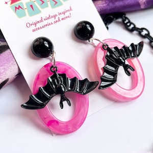 May include: Pink and black bat earrings with a black bead at the top. The earrings are on a white background with a pink and black chain in the foreground. The text "Mid Century Missy Original vintage inspired accessories and more!" is on a white card behind the earrings.