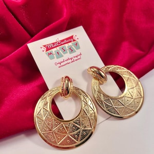 May include: A pair of gold-toned, vintage-inspired earrings. Each earring features a large, circular design with geometric patterns and a smaller, decorative top. The earrings are displayed on a white card with the text "Mid-Century Missy" and "Original vintage inspired accessories and more!"