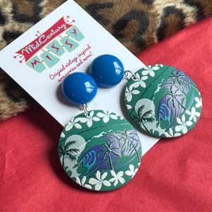 May include: A pair of blue and green dangle earrings with a tropical print and white flowers. The earrings are made of polymer clay and feature a blue cabochon.