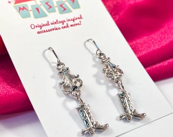 Pode incluir: Brincos prateados com um design decorativo e um berloque de bota de cowboy. Os brincos são exibidos em um cartão branco com o texto "MidCentury Missy" e "Original vintage inspired accessories and more!"