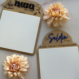 Könnte beinhalten: Zwei Holz-Spielabend-Anzeigetafeln mit weißen Schreibflächen. Auf einer steht "The Freezer Book Club Trivia" mit einer Geistergrafik. Auf der anderen steht "The Saldin Family Game Night Scoreboard". Zwei pfirsichfarbene Kunstblumen sind ebenfalls im Bild.