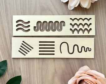 May include: Two light-colored wooden boards with various line designs cut out. The top board features wavy lines and a zig-zag pattern. The bottom board has diagonal lines, horizontal lines, and a curved line design. Decorative flowers are in the background.