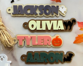 May include: Wooden name tags with layered acrylic lettering. The names "Jackson", "Olivia", "Tyler", and "Aaron" are displayed in various colors. Halloween-themed accents include a ghost, cat, pumpkin, and bat.