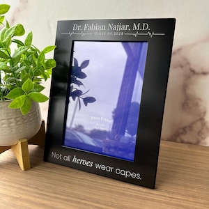 May include: A black photo frame with the text "Dr. Fabian Najjar, M.D. CLASS OF 2025" and "Not all heroes wear capes." The frame is designed to hold a 5" x 7" (13 x 18cm) photo. A small plant in a white pot is visible in the background.