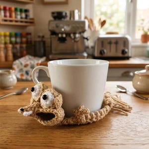 May include: A cream-coloured ceramic mug with a crocheted brown creature wrapped around it. The creature has large black eyes and an open mouth. The mug rests on a wooden table, with kitchen appliances visible in the background.