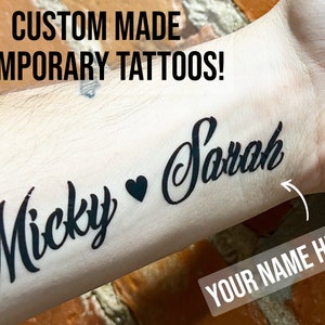 Pu&ograve; includere: Tatuaggi temporanei personalizzati con i nomi "Micky" e "Sarah" sul braccio di una persona. Il testo "YOUR NAME HERE" &egrave; anche visibile.