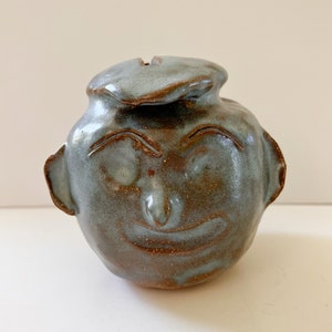 May include: A ceramic money box shaped like a face with a smiling expression. The money box is glazed in a light blue and brown colour.