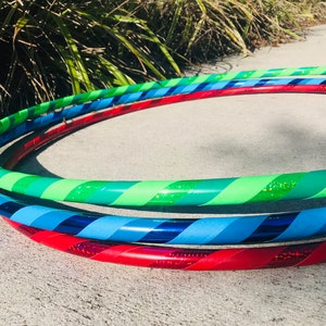 May include: Three colorful hula hoops with striped patterns. The top hoop is green and green glitter, the middle hoop is blue and black, and the bottom hoop is red and red glitter.