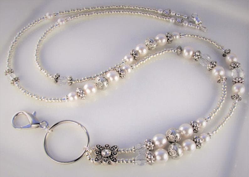 May include: Elegant silver lanyard with a double strand design. The lanyard features white faux pearls, clear crystal beads, and decorative silver accents. A silver clasp and key ring are attached at the bottom. The lanyard is approximately 30 inches long.