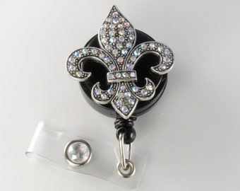 May include: A black retractable badge reel with a silver fleur-de-lis design. The fleur-de-lis is encrusted with iridescent rhinestones. A clear plastic strap and a silver-colored metal clip are attached to the reel.