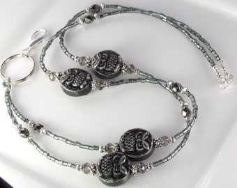 May include: A beaded lanyard with a silver clasp and key ring. The lanyard features dark gray beads and decorative, round, dark gray beads with a floral design. The lanyard is designed to be worn around the neck.