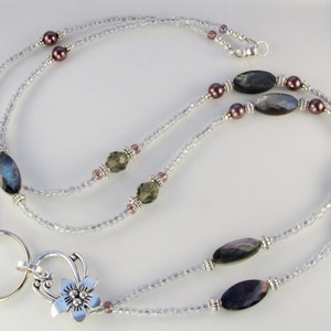 May include: A beaded lanyard with a silver-tone flower charm and key ring. The lanyard features clear seed beads, iridescent oval beads, and small purple beads. The lanyard is designed to be worn around the neck.