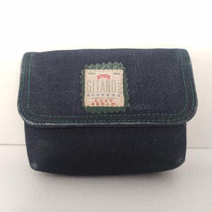 May include: A dark blue denim pouch with a rectangular flap and green stitching along the edges. Centered on the flap is a beige fabric patch with the text "TRADE MARK GITANO EXPRESS AGED DENIM" printed in red and black ink with a jagged green border.