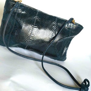 May include: A dark teal clutch with a snakeskin pattern. The bag has a long, thin strap and gold-colored hardware. The texture of the bag is visible, and the strap is knotted at the end.