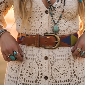 Op de afbeelding: Crèmekleurige gehaakte jurk met een bruine leren riem met een messing gesp en kleurrijke geometrische accenten. De outfit is gecombineerd met turkooiskleurige sieraden en meerdere gouden armbanden en ringen.