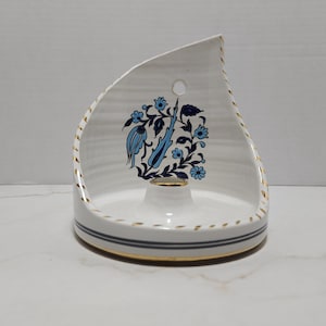 May include: A white ceramic candle holder with a curved back and a central circular base. The piece features a blue floral pattern, gold-painted edges, and a thin gray horizontal stripe around the base. A small hole is located near the top edge.