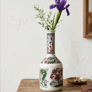 May include: White ceramic vase with a colorful floral pattern, holding a purple iris and greenery. The vase has a narrow neck and a wider base, with red, green, and blue floral designs. The vase is on a wooden table.