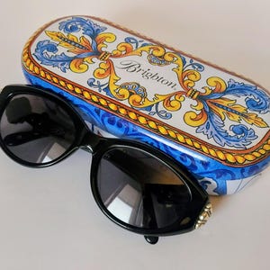 Puede incluir: Gafas de sol negras con un acento decorativo de pedrería en la montura, junto a un estuche a juego. El estuche es blanco con un diseño floral azul y amarillo y la palabra "Brighton".