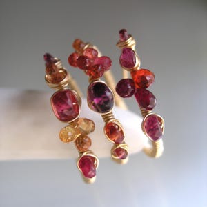 May include: Three wire-wrapped rings featuring clusters of faceted gemstones in shades of red, orange, pink, and deep purple. Each ring is constructed from thin gold-toned wire that secures the various small, polished, and irregular-shaped stones.