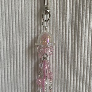 Könnte beinhalten: Ein rosa Quallen-Schlüsselanhänger mit einer transparenten, irisierenden Kuppel und einer rosa Schleife. Der Schlüsselanhänger hat hängende rosa Perlen und silberfarbene Ketten, die an einem silbernen Verschluss und einer dünnen Silberkette befestigt sind.