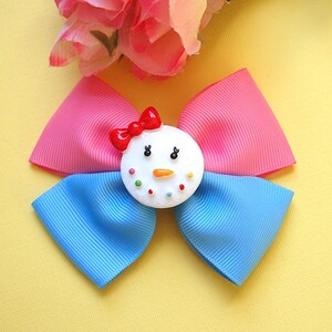 Peut inclure: Un nœud pour cheveux fait à la main avec un motif de bonhomme de neige. Le nœud a des rubans roses et bleus, avec un nœud rouge sur le dessus du bonhomme de neige. Le bonhomme de neige a un visage blanc avec des yeux noirs, un nez en carotte et des points colorés. Le fond est jaune.
