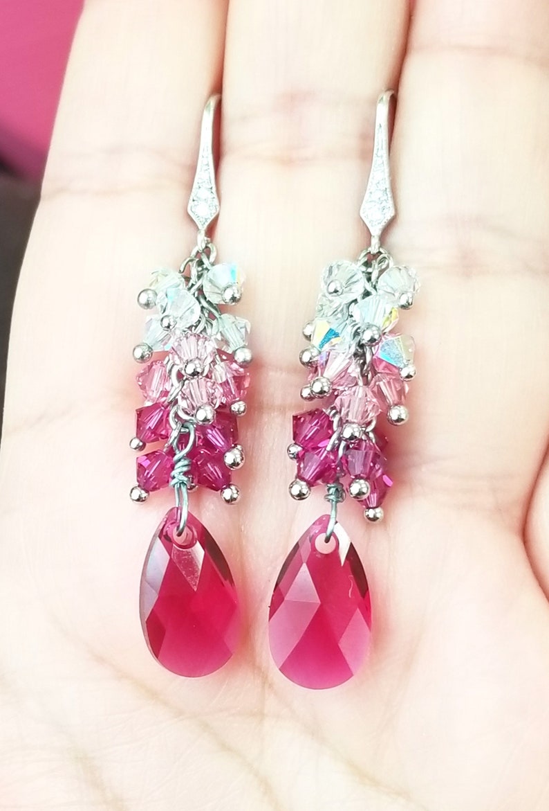 Peut inclure: Boucles d'oreilles pendantes argent&eacute;es avec un cristal rouge en forme de larme et une cascade de petits cristaux dans des tons de rose, blanc et iris&eacute;.