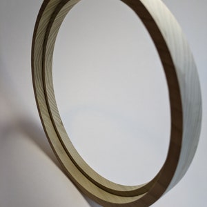 May include: A circular wooden frame with a light-colored wood grain. The frame is angled, casting a shadow on the white background. The design is simple and modern, with a focus on the natural wood texture.