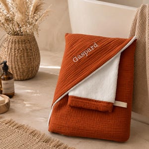 Peut inclure: Une serviette à capuche pliée de couleur rouille avec une doublure intérieure blanche et un liseré blanc. Le prénom Gaspard est brodé en fil blanc sur le tissu extérieur rouille. La serviette présente un tissage en coton mousseline texturé.