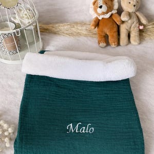 Peut inclure: Une couverture de bébé vert foncé avec une bordure blanche et moelleuse. Le nom "Malo" est brodé en écriture blanche sur la couverture. Deux jouets en peluche sont en arrière-plan.