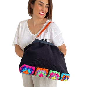 May include: A black crocheted tote bag with a colorful granny square trim. The bag has a top closure with a metal clasp and a brown leather strap.