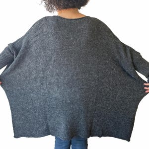 May include: A dark gray, oversized knit sweater with a wide, flowing silhouette. The sweater has long sleeves and a ribbed knit texture on the cuffs. The model is wearing blue jeans.
