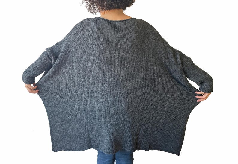 May include: A dark gray, oversized knit sweater with a wide, flowing silhouette. The sweater has long sleeves and a ribbed knit texture on the cuffs. The model is wearing blue jeans.