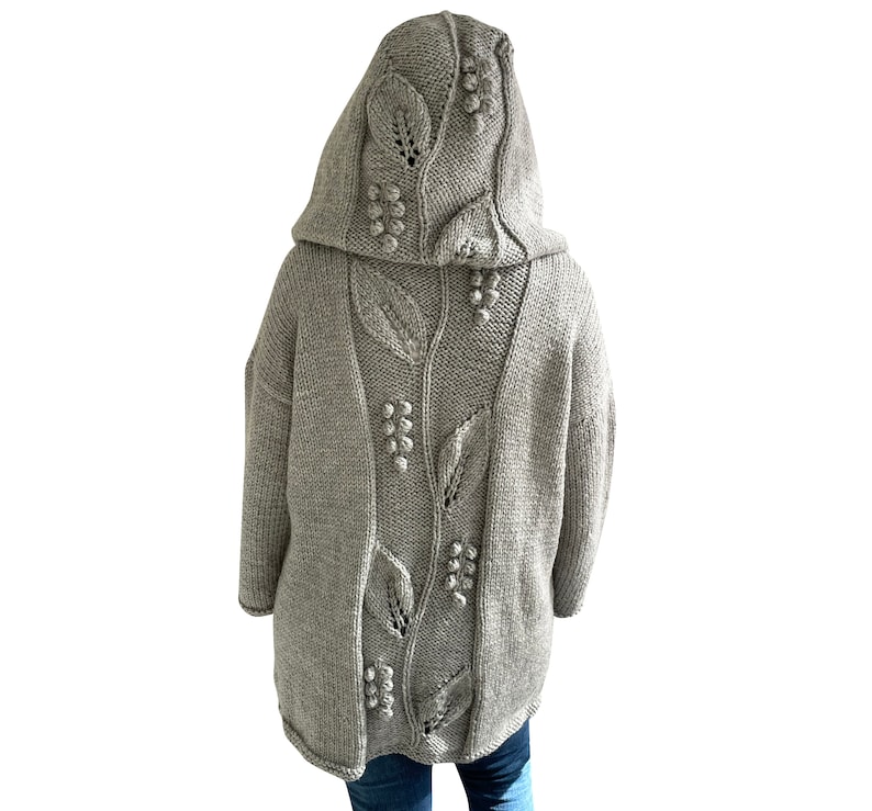K&ouml;nnte beinhalten: Ein grauer Kapuzenpullover mit einem in den R&uuml;cken gestrickten Blatt- und Beerenmuster. Der Pullover ist aus weichem, dickem Garn gefertigt.