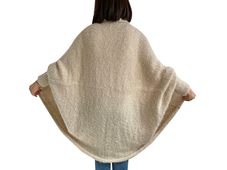 Puede incluir: Un c&aacute;rdigan de punto de color crema con un corte holgado y fluido. El c&aacute;rdigan tiene un cuello alto y mangas largas.