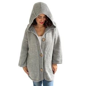 K&ouml;nnte beinhalten: Ein grauer Kapuzenpullover mit Kn&ouml;pfen. Der Pullover ist gestrickt und hat eine lockere Passform.