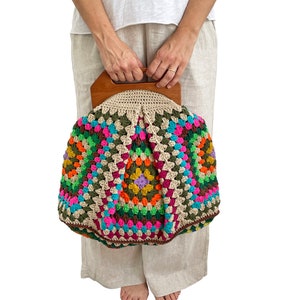 May include: A colorful, handmade crochet bag with a wooden handle. The bag features a vibrant patchwork design with squares of pink, green, orange, and purple. The top of the bag is a neutral beige color.