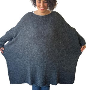 May include: A dark gray, oversized knit sweater with long sleeves. The sweater has a wide, boat neckline and a loose, flowing silhouette. The fabric appears to be a soft, textured knit, perfect for cooler weather. The model is wearing blue jeans.