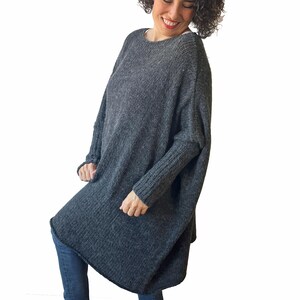 May include: A dark gray, oversized knit sweater with a loose, flowing design. The sweater has long sleeves and a rounded neckline. The fabric appears to be a soft, textured knit. The sweater is worn with blue jeans.