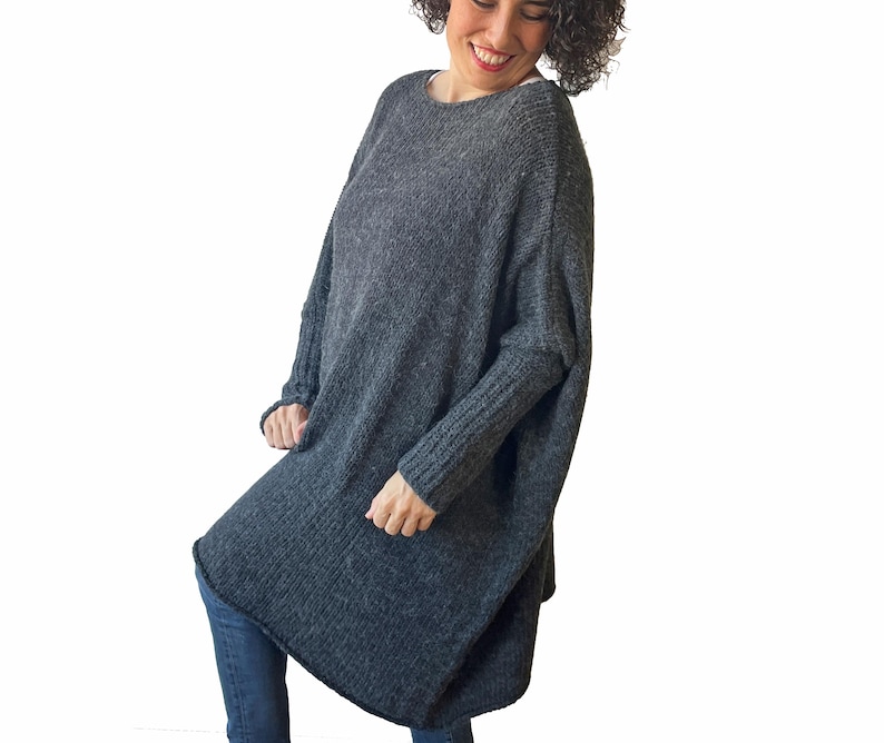 May include: A dark gray, oversized knit sweater with a loose, flowing design. The sweater has long sleeves and a rounded neckline. The fabric appears to be a soft, textured knit. The sweater is worn with blue jeans.