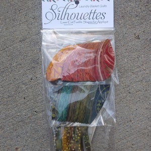 May include: A clear plastic bag containing laser-cut fusible shapes for appliqué. The shapes are in shades of orange, red, green, and blue. The bag has a white label with the word "Silhouettes" and the text "Laundry Basket Quilts".
