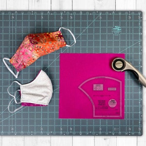May include: Two fabric face masks with elastic ear loops, one with a colorful floral pattern and the other with a silver and pink design, lie on a gray cutting mat. A pink fabric square, a rotary cutter, and a face mask template are also present.