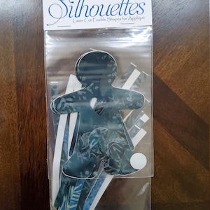 May include: A package of laser-cut fusible shapes for appliqué. The package includes various shapes, including a silhouette of a person. The shapes are in shades of blue and white, with a patterned design. The package is labeled "Snowbird Silhouettes."