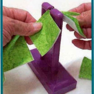 May include: A purple craft tool with a rectangular base and vertical supports. Green patterned fabric squares are being inserted into the tool. The image shows the process of creating a craft project.