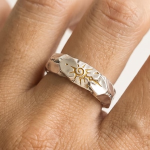 May include: A silver-toned ring featuring a textured, uneven surface and a central gold-coloured sun motif with a spiral centre and radiating lines. The band has a rugged, hammered appearance with distinct facets and a polished metallic finish.
