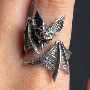Puede incluir: Un anillo de metal plateado con un diseño detallado de murciélago. La cabeza del murciélago está centrada con orejas puntiagudas y alas texturizadas que envuelven el dedo, creando un estilo de banda abierta con un acabado pulido y oxidado.