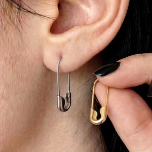 May include: A pair of safety pin earrings, one silver and one gold, are shown. The silver earring is in the ear, while a hand holds the gold earring. The earrings are simple and minimalist, with a classic safety pin design.