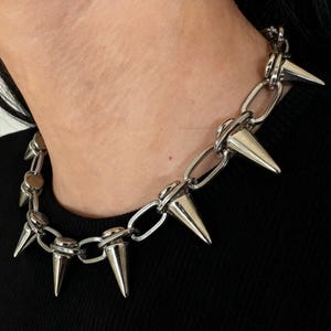 May include: A silver-colored chain necklace with large, pointed spikes. The necklace is made of metal and has a chunky chain design. The spikes are evenly spaced around the collar, creating a bold, edgy look.