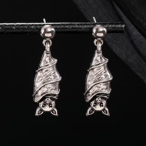 May include: A pair of silver-toned drop earrings featuring a detailed bat design. Each earring depicts a bat hanging upside down with textured wings and visible facial features, suspended from a small polished silver-toned ball stud post.