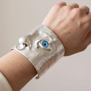 May include: A wide silver cuff bracelet featuring a sculpted human nose and a single realistic blue eye. The metal surface has a hammered texture. A small beaded ring hangs from the nose, and two tiny silver spheres are positioned near the eye.