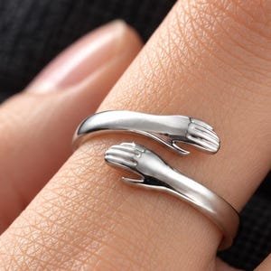 Könnte beinhalten: Ein silberfarbener offener Ring mit zwei Händen, die sich um den Finger legen und ein Umarmungsdesign bilden. Das Band ist glatt und poliert, wobei sich die Hände an der Vorderseite treffen und eine durchgehende, verstellbare Schlaufe bilden.